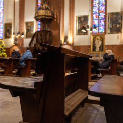 A photo showing two empty church pews in a catholic church. In the background, a column of pews with some people sat among them is visible, though blurred. The church is richly decorated with stain glass windows in the background.