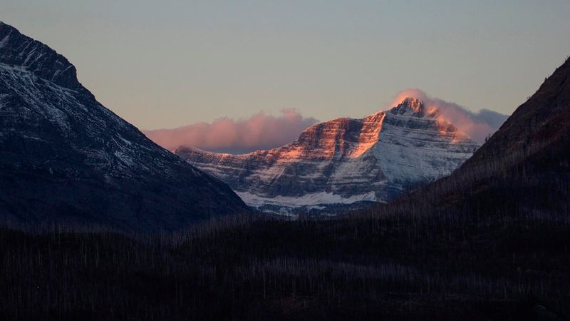 A photo showing the mountain in the distance, framed between the silhouettes of two closer mountains. Triple Divide Peak is bathed in the soft reddish light of an early morning, while the rest of the landscape is shadowy. The sky surrounding it is clear, with some low reddish/purple clouds gathering just behind it.