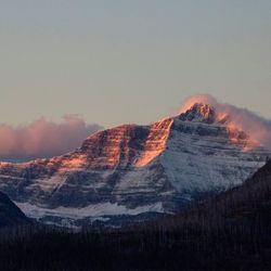 A photo showing the mountain in the distance, framed between the silhouettes of two closer mountains. Triple Divide Peak is bathed in the soft reddish light of an early morning, while the rest of the landscape is shadowy. The sky surrounding it is clear, with some low reddish/purple clouds gathering just behind it.